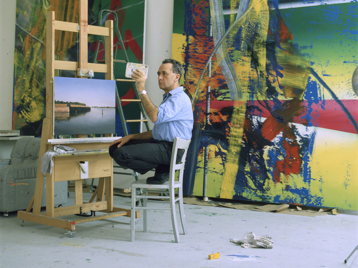 Richter squats on a metal chair in his art studio, in front of an easel displaying a realistically painted landscape, with two towering abstract paintings leaning on the floor against the wall behind him. He holds and looks at a small compact mirror which presumably reflects the landscape painting on the easel next to him.